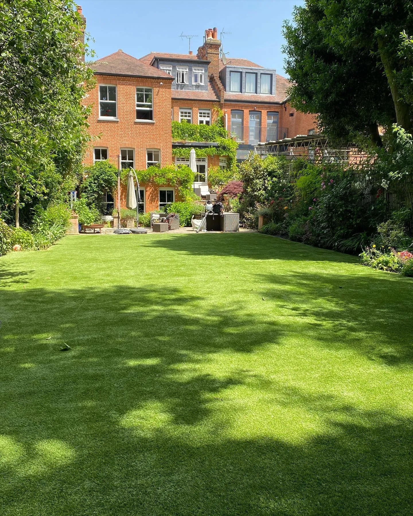 A large artificial lawn in a mature garden behind a red brick house